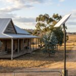 Photo of a Starlink satellite dish mounted on a pole next to a modern Australian rural farmhouse with weathered timber siding, corrugated metal roof, eucalyptus trees and rolling pastoral hills